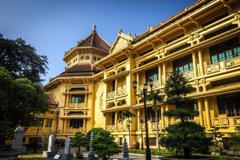 Yellow colonial-style building with ornate balconies and a central tower, set in a sunny courtyard with trees and lampposts.